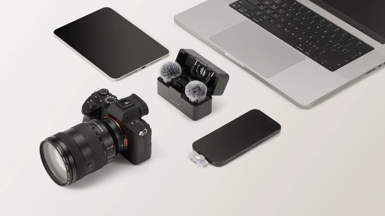 Open DJI Mic Mini 2S charging case with two transmitters, next to a camera, a phone with the receiver, a tablet and a laptop