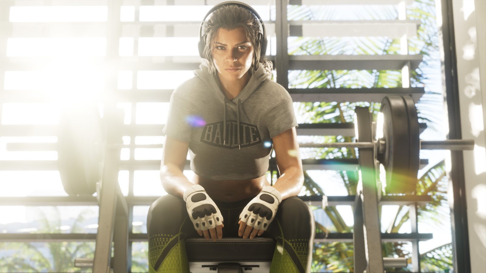 Lucia Caminos sentada en un banco de gimnasio con cascos, sudadera corta gris con la palabra Baddie y guantes de entrenamiento